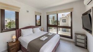 A modern hotel room with a large bed, a TV, and a balcony with a view of buildings and trees. - Ayia Napa Seaside Villa Ionion H15 (Sotira)