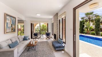 Una sala moderna con un sofá, sillones, una mesa de centro y una chimenea. Hay un ventanal con vista a una piscina y palmeras.
