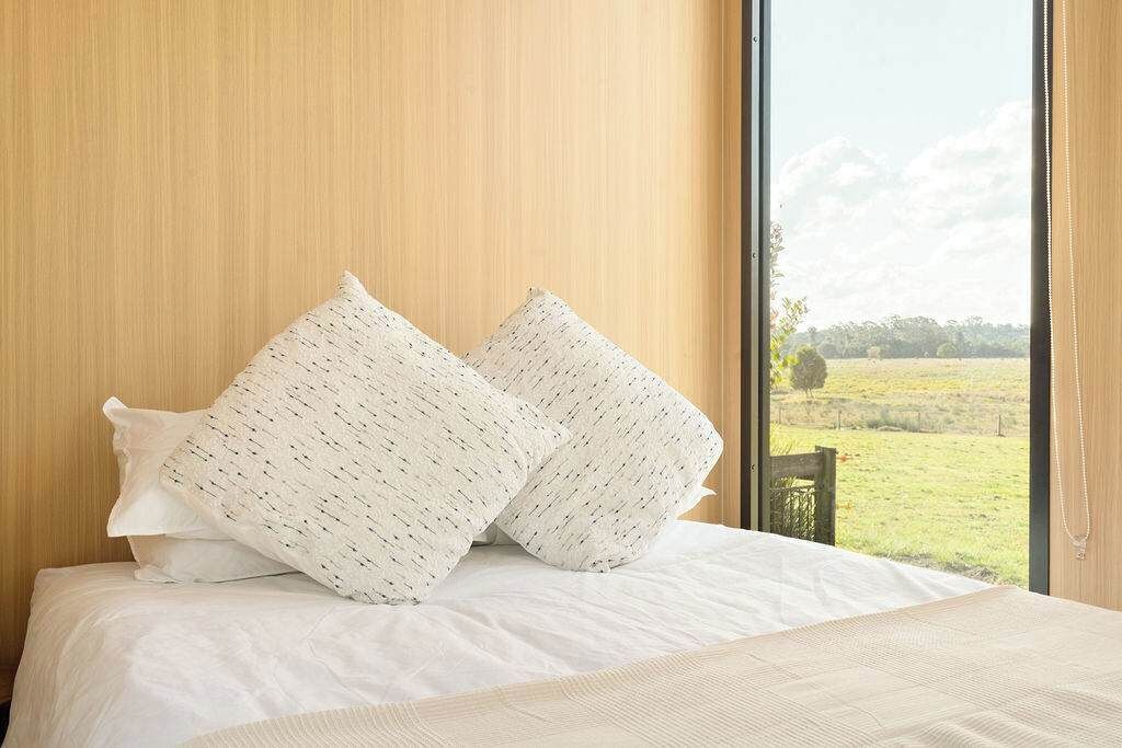 A bed with white bedding and two patterned pillows, a wooden wall, and a view of a green field through a window.
