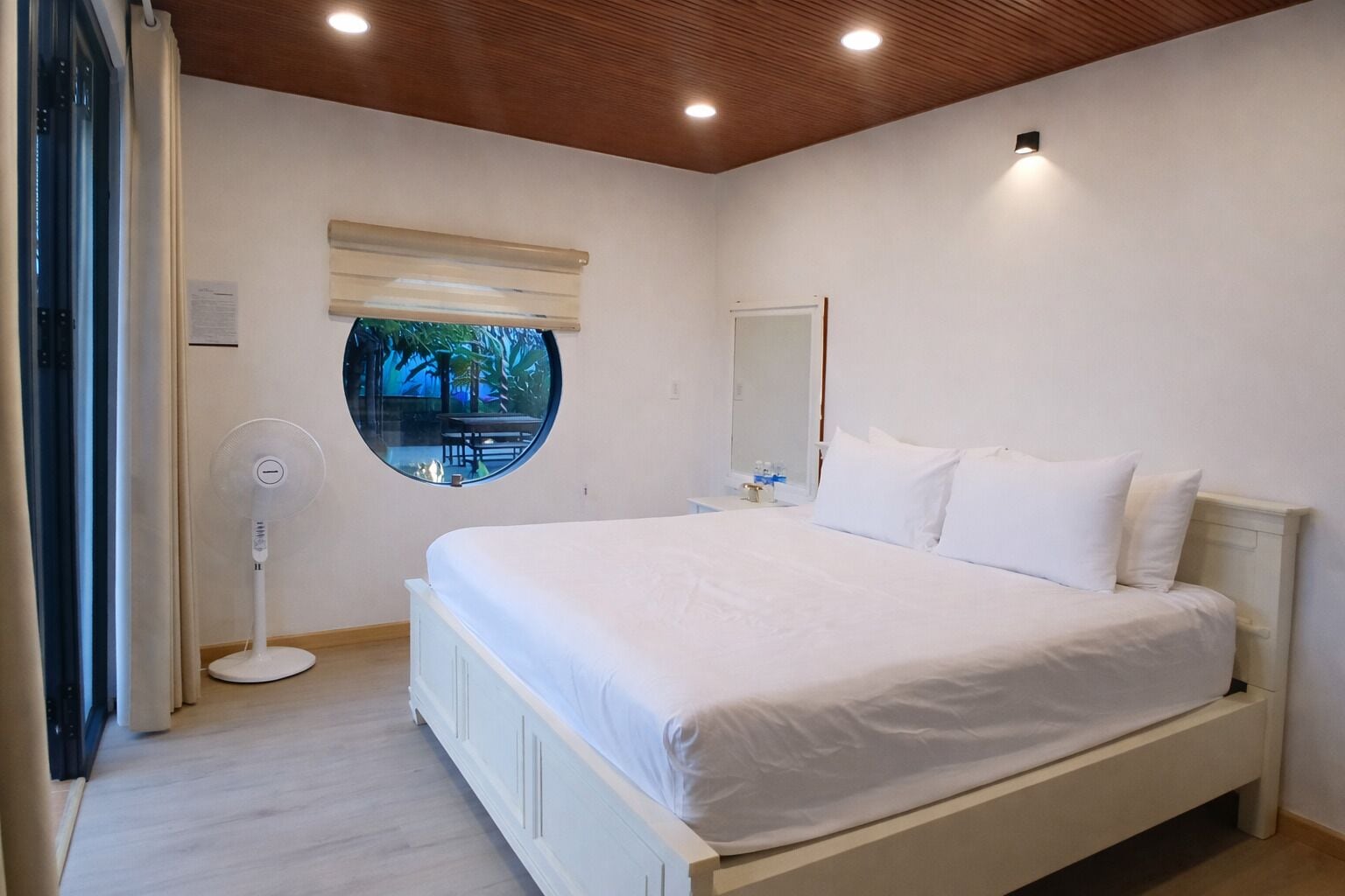 A bedroom with a bed, a round mirror, a ceiling fan, and a window with a view of palm trees.