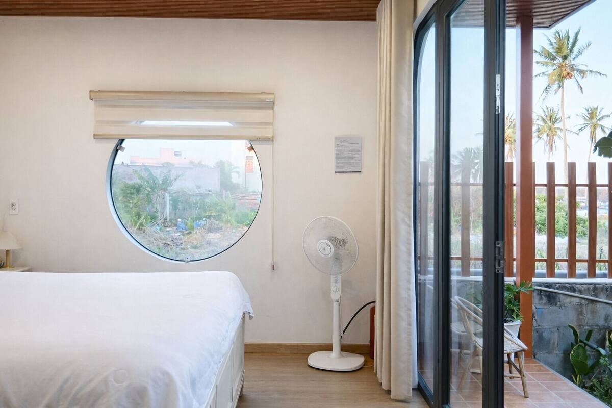 Une chambre avec un lit, une fenêtre ronde, un ventilateur sur pied et un balcon agrémenté de plantes.