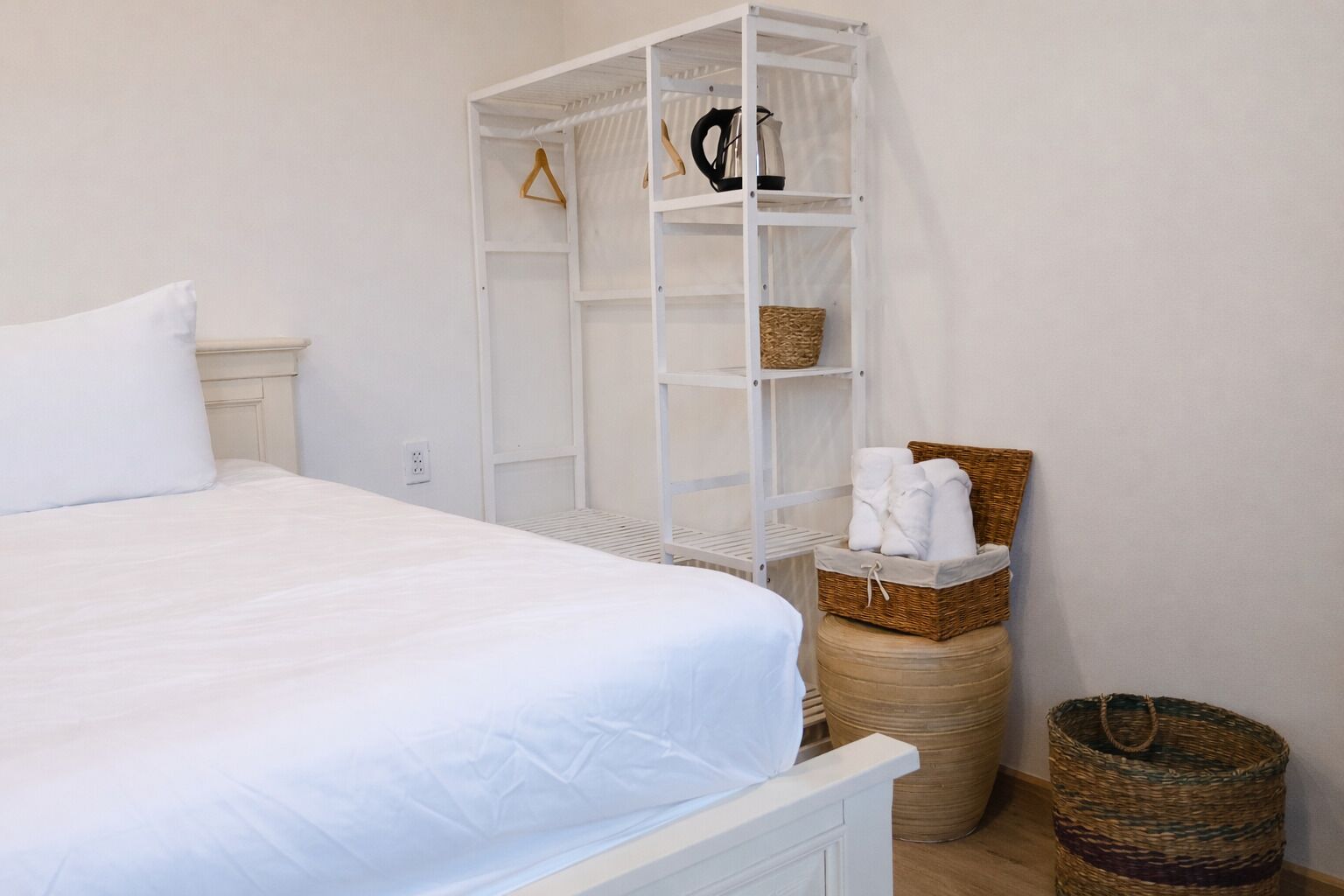 Una cama bien tendida con sábanas blancas, un armario blanco con espacio para colgar y una cesta de mimbre con toallas en el suelo.