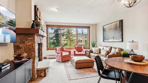 A modern living room with a stone fireplace, a large window with a view of greenery, a dining area with a round table, and a cozy seating arrangement.