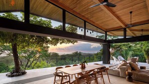 A spacious outdoor dining area with a wooden table and chairs, a sofa, and a view of lush greenery.