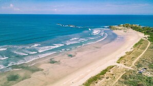 An aerial view of a beach with clear blue waters, sandy shore, and sparse vegetation.
