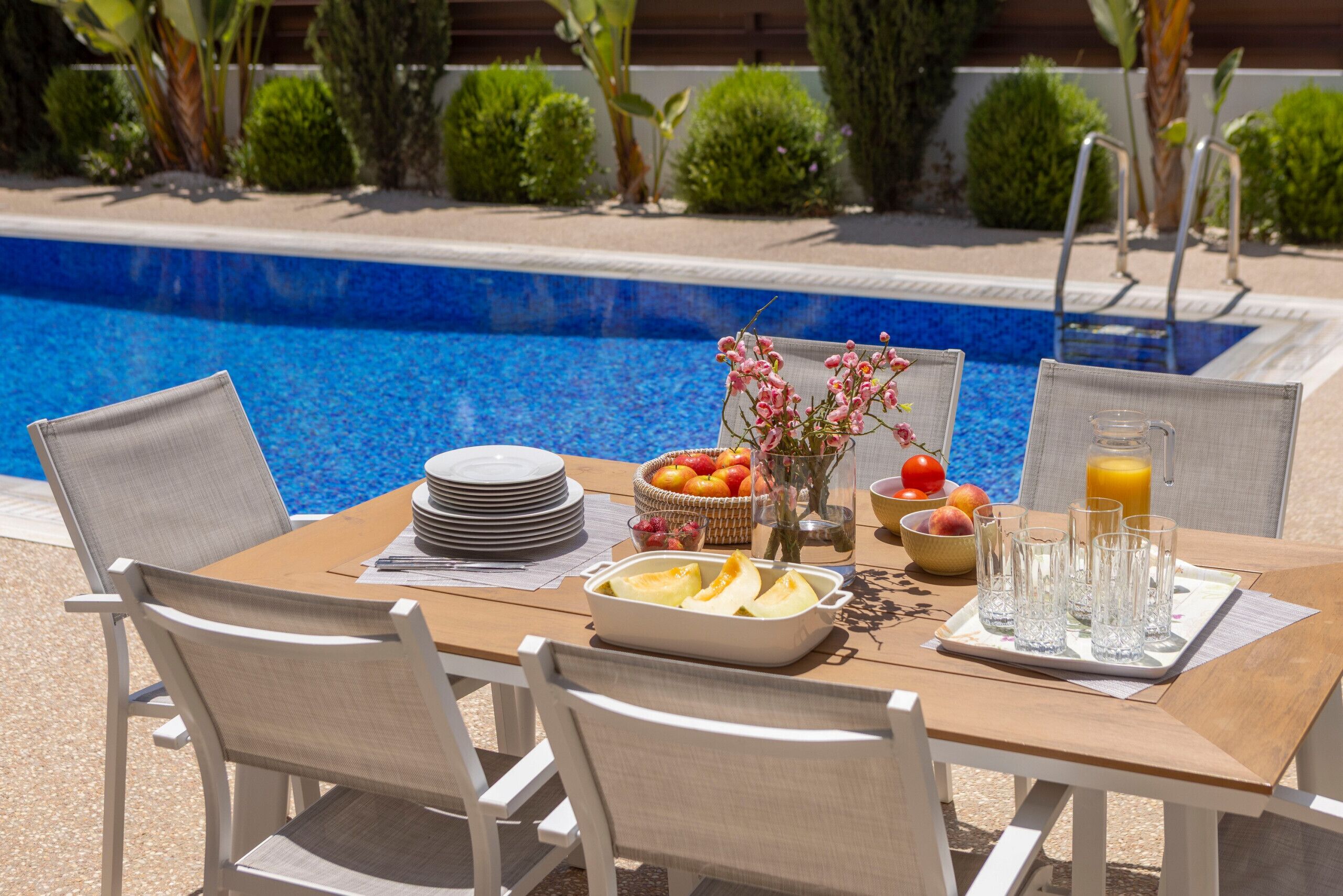 Uma mesa de jantar com cadeiras, pratos, uma cesta de frutas, um vaso com flores e copos, além de uma jarra de suco de laranja perto de uma piscina.