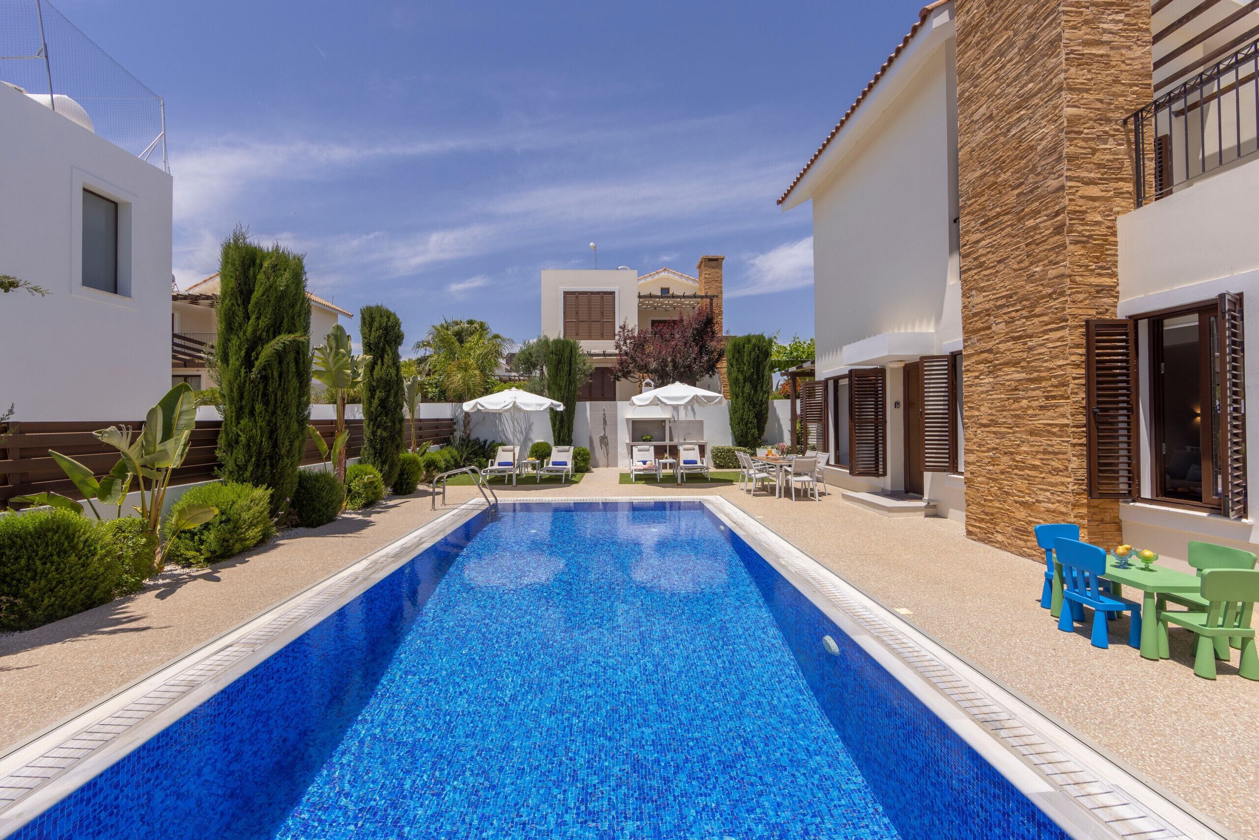 Piscina con sillones de descanso y sombrillas, rodeada de follaje y edificios modernos.
