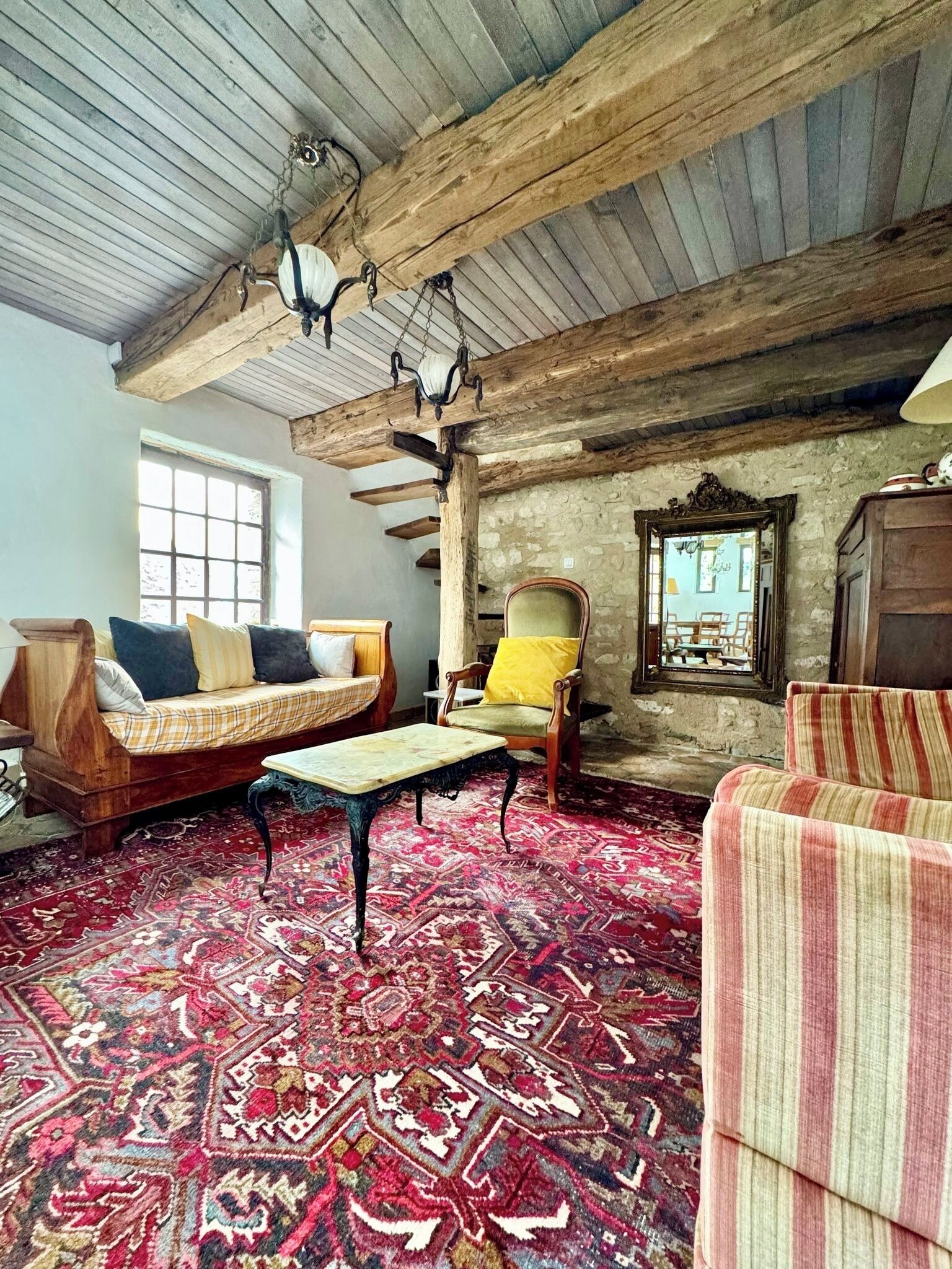 Una habitación rústica con vigas de madera a la vista, alfombra estampada, sofá, sillón y una mesa.