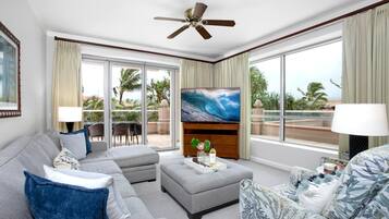 A spacious living room with a large flat-screen TV, a ceiling fan, and a view of palm trees through large sliding glass doors.