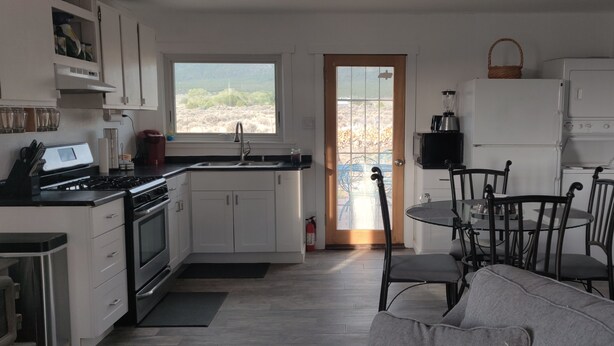 A modern kitchen with white cabinets, a stove, a sink, and a dining area with a table and chairs. - Hidden Haven: Open Concept Inside, Dramatic Views Outside (Questa)