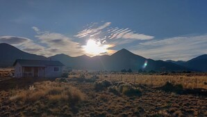 A small house with a fenced yard, surrounded by mountains and a clear sky with the sun setting. - Hidden Haven: Open Concept Inside, Dramatic Views Outside (Questa)