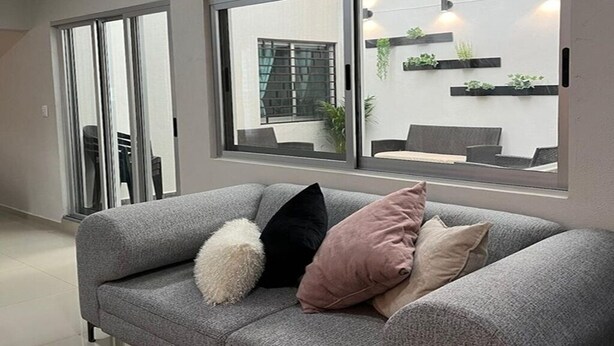 Un salon moderne avec un canapé gris, un coussin blanc moelleux et un coussin rose. De grandes fenêtres offrent une vue sur un espace extérieur agrémenté de plantes et d’un coin salon.