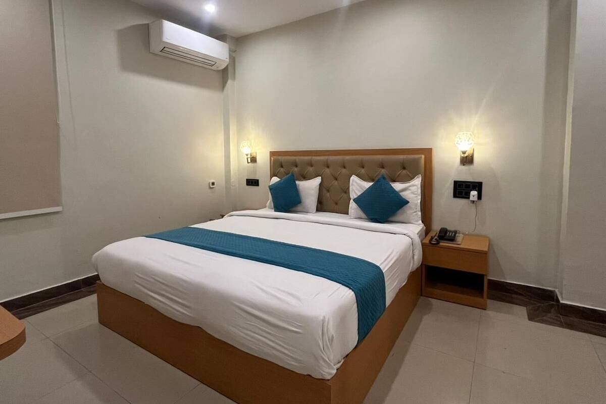 A hotel room with a large bed, a bedside table, an air conditioning unit, and wall-mounted lighting.