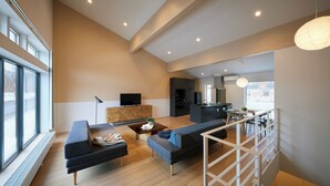 Ein geräumiger Wohnbereich mit einer Couch, einem Couchtisch und einem Fernseher auf einem Holzkasten. Es gibt eine Küchenzeile mit einem Esstisch und Stühlen. Der Raum verfügt über große Fenster und eine Treppe mit Geländer.