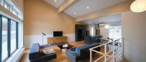Amplia sala de estar con sofá, mesa de centro y televisor sobre un mueble de madera. Zona de cocina con mesa y sillas. La habitación tiene ventanas grandes y una escalera con barandilla.