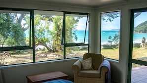 A bedroom with a bed, a chair, and a small table. There are large windows with a view of a beach and trees.