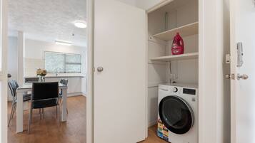 A laundry room with a washing machine, a shelf with detergent, and a door leading to a kitchen area.