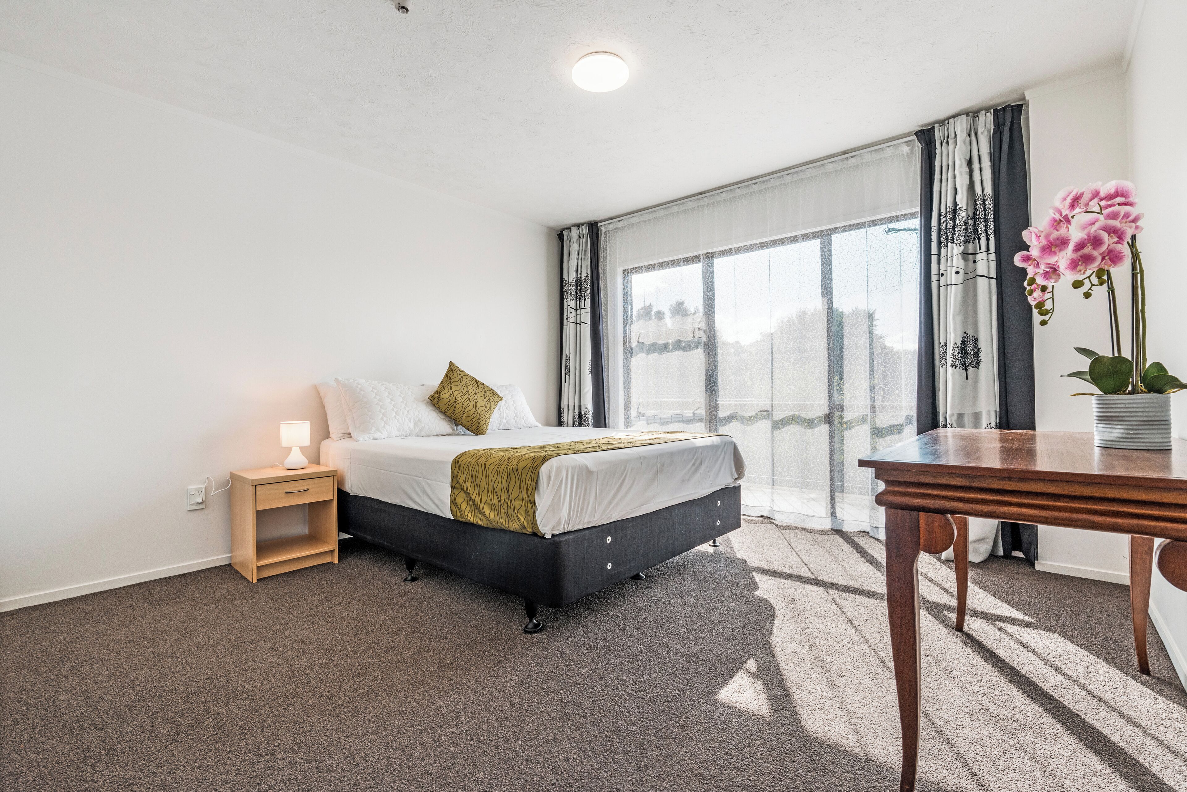 Quarto com cama, mesa de cabeceira, abajur e vista para o exterior pela janela.