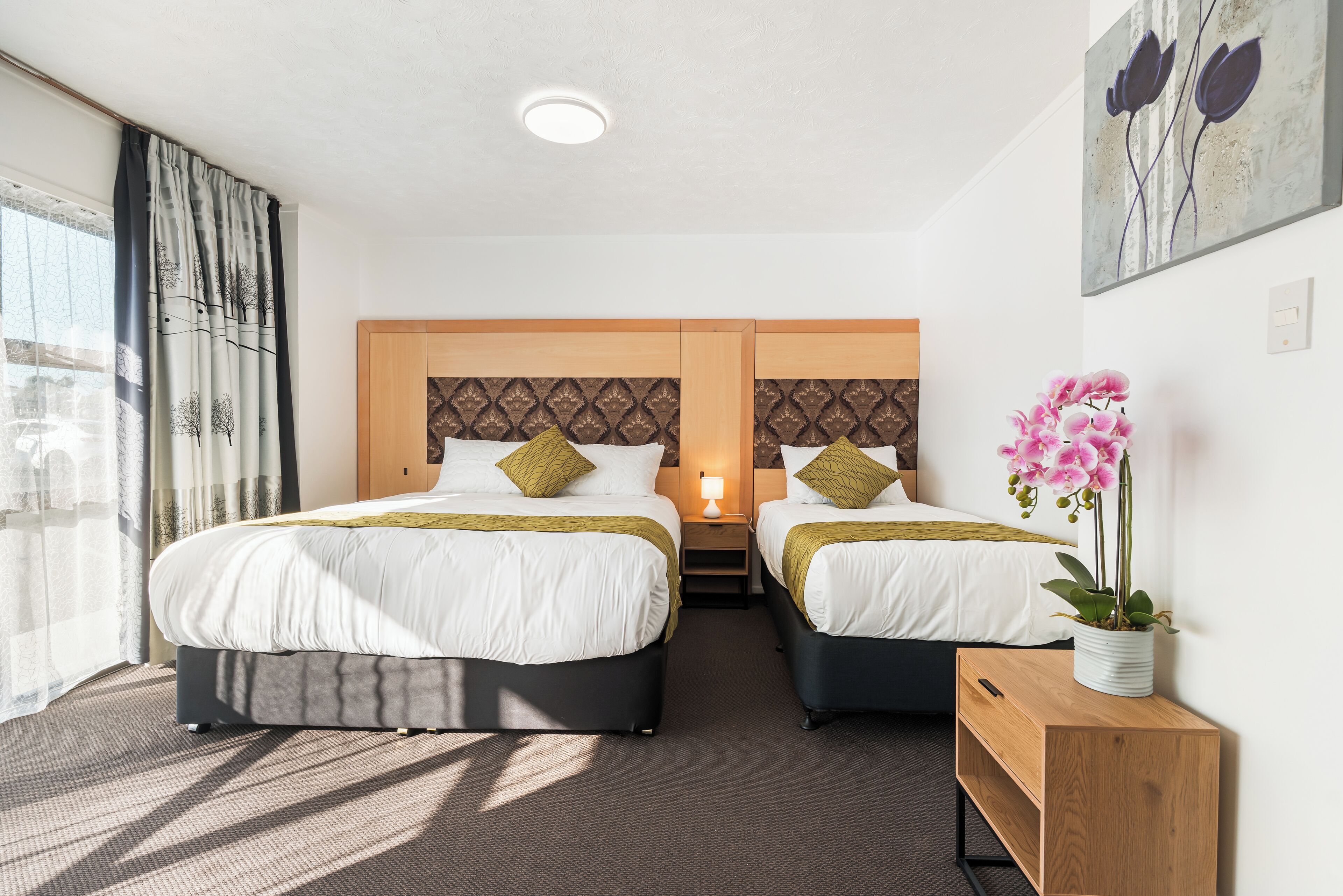 A hotel room with two beds, a nightstand, a lamp, a wooden side table, and a potted orchid.