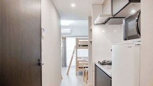 Una cocina estrecha con refrigerador, estufa y microondas. Una escalera de madera está apoyada contra la pared, y hay una puerta abierta al final del pasillo.