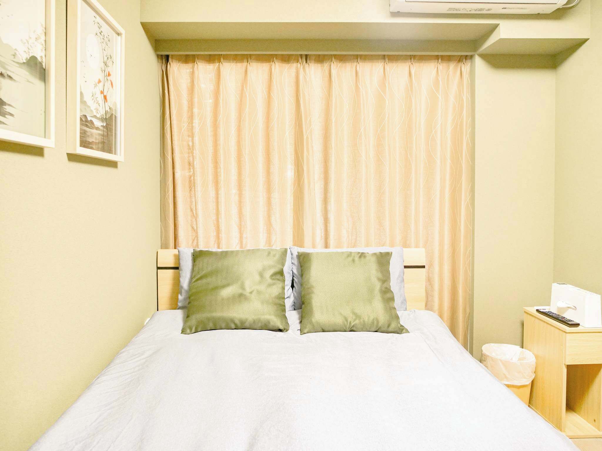 Een net opgemaakt bed met twee kussens, een nachtkastje met een afstandsbediening en zakdoos, en een wandairconditioner.
