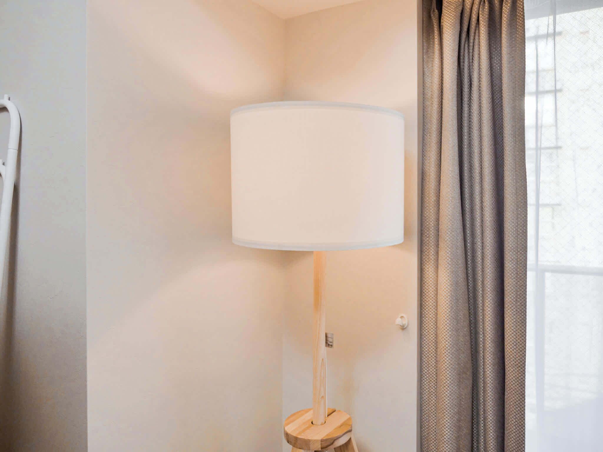 Eine Stehlampe mit weißem Lampenschirm auf einem Holzfuß, neben einem Fenster mit Vorhang.