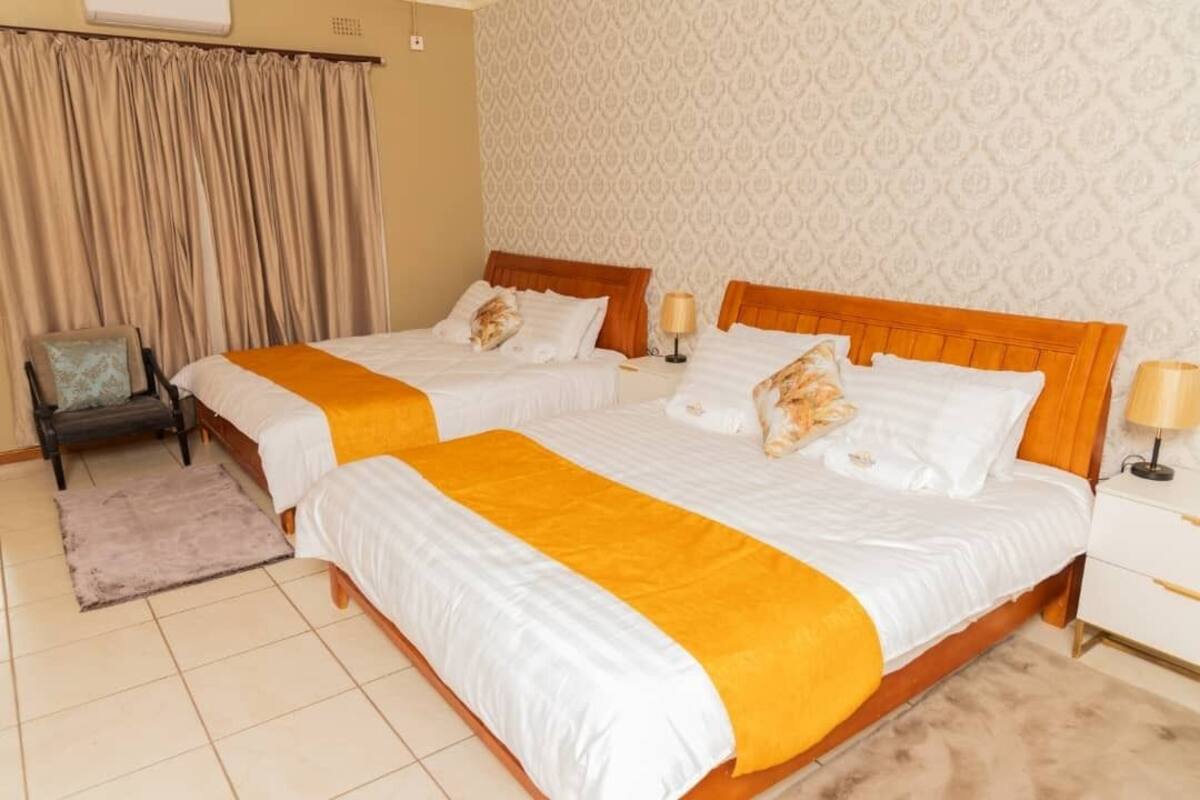 A hotel room with two beds, beige curtains, patterned wallpaper, and a small chair.