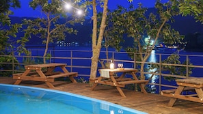 Un espace aménagé au bord de la piscine, avec des tables de pique-nique en bois et une vue sur une étendue d’eau la nuit.