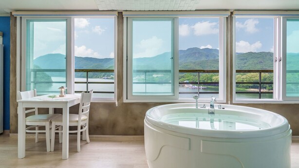A room with a large bathtub, a dining table with chairs, and large windows with a view of mountains and a lake. - Gapyeong Nami Island Pool & Spa Pension (Gapyeong)
