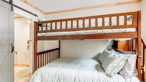Uma cama beliche com estrutura de madeira e roupa de cama branca. Há uma porta à esquerda e um pequeno prateleira embaixo da cama.