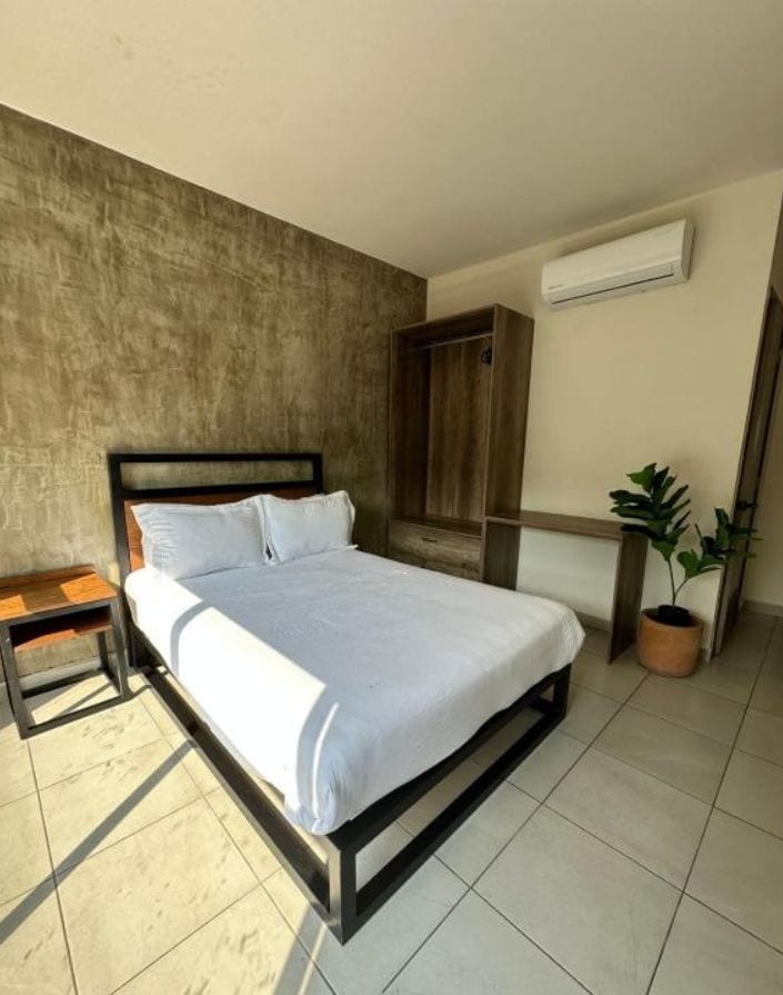 A bedroom with a bed, bedside table, potted plant, and an air conditioning unit.
