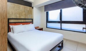 A modern hotel room with a large bed, a wooden headboard, a window with blinds, and a small bedside table. - Rau Departamentos (Guadalajara)