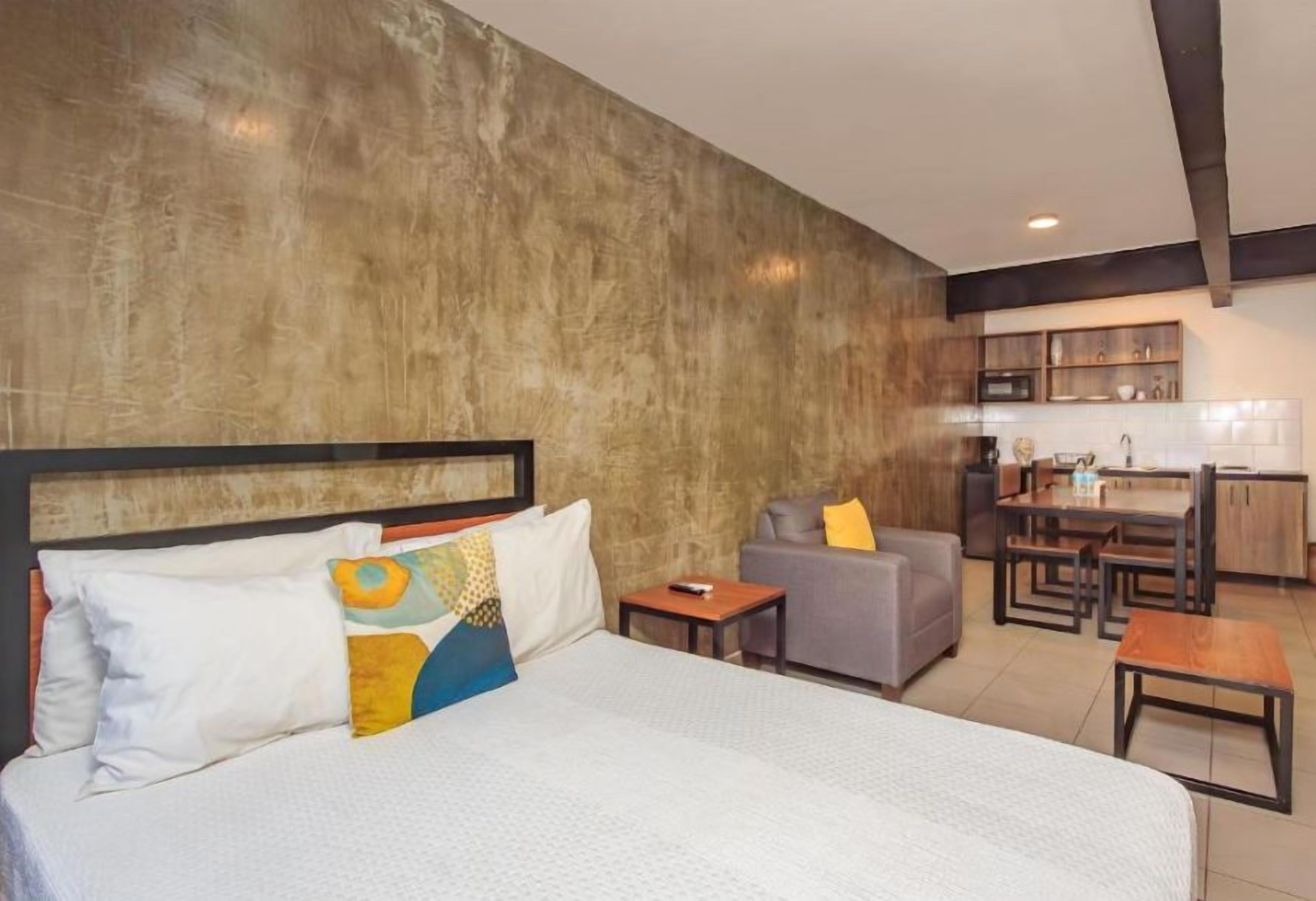 A hotel room with a large bed, a sofa, a small table, and a kitchenette in the background.