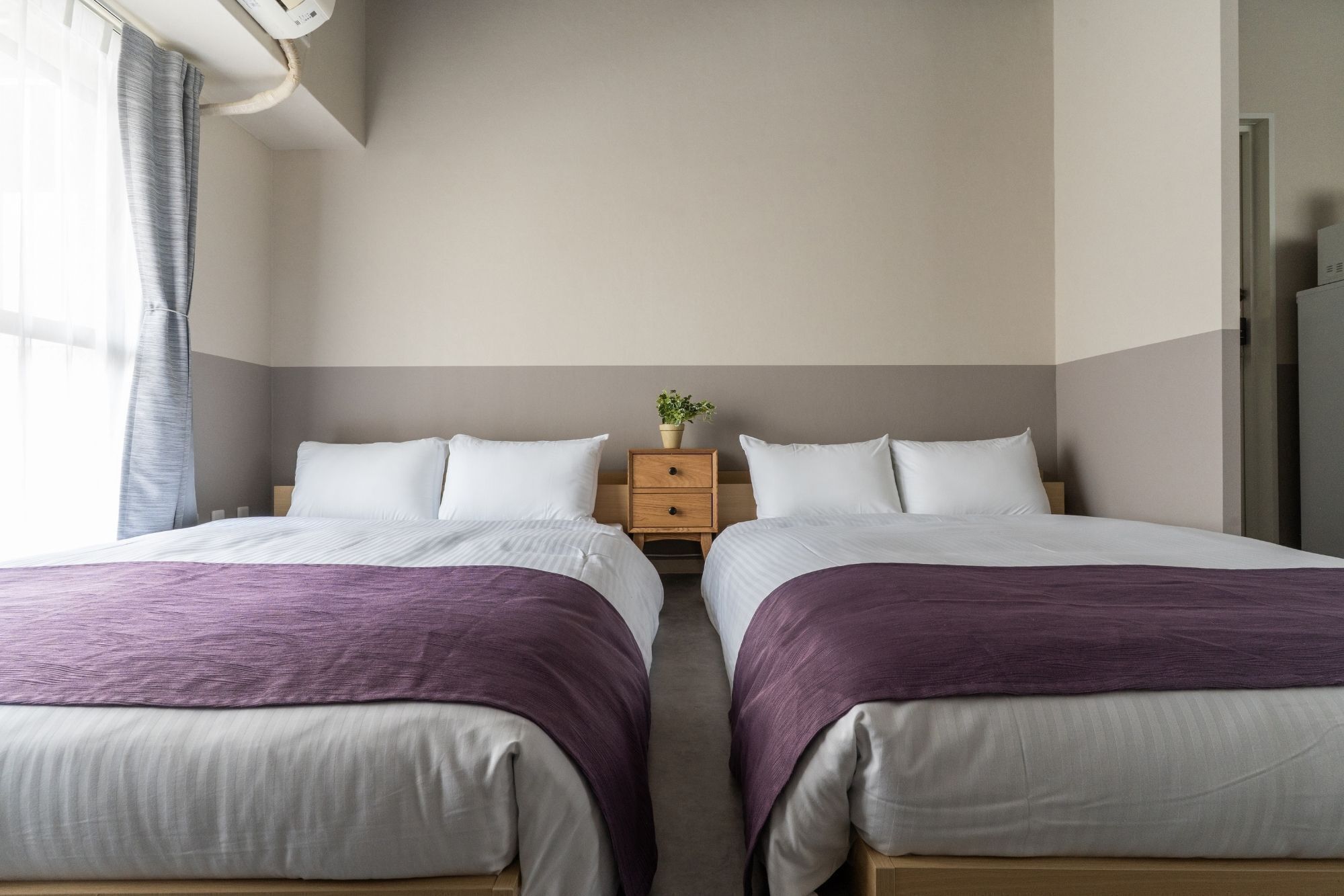 A hotel room with two beds, a nightstand with a plant, and a view of a staircase.