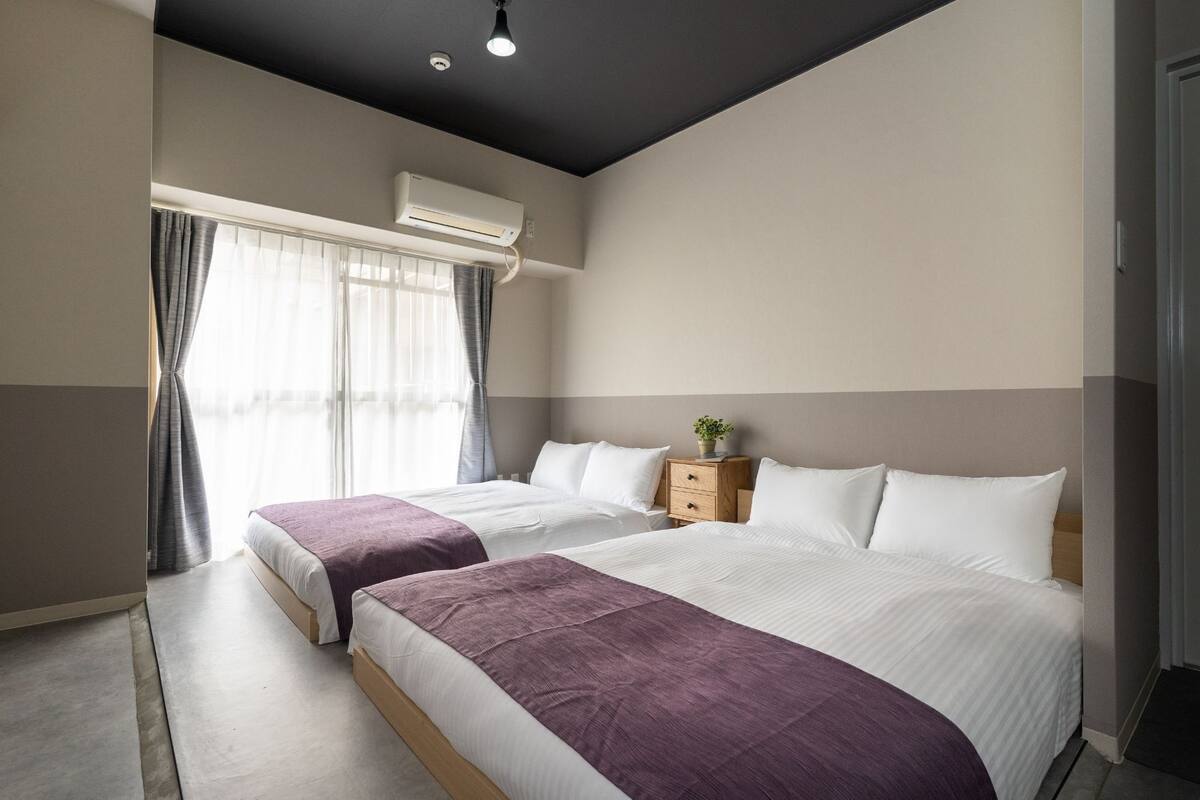 Hotellihuone, jossa on kaksi sänkyä, pieni puinen yöpöytä, ruukkukasvi ja ikkuna, jossa on verhot.