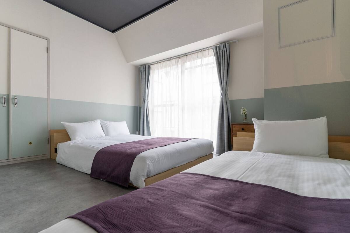 Hotellihuone, jossa on kaksi sänkyä, vaatekaappi, ikkuna verhoilla ja pieni yöpöytä, jossa on maljakko.