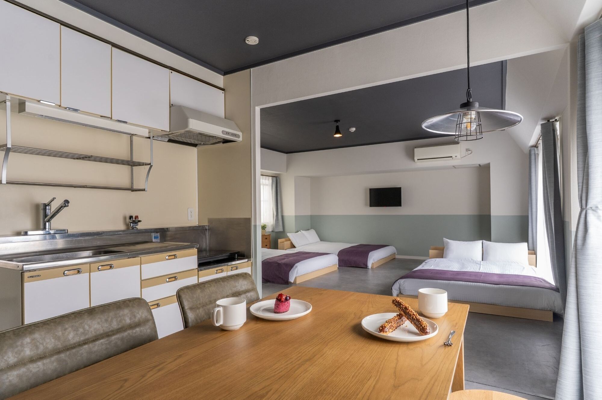 A modern hotel room with a kitchenette, two beds, a dining table, and a large window.