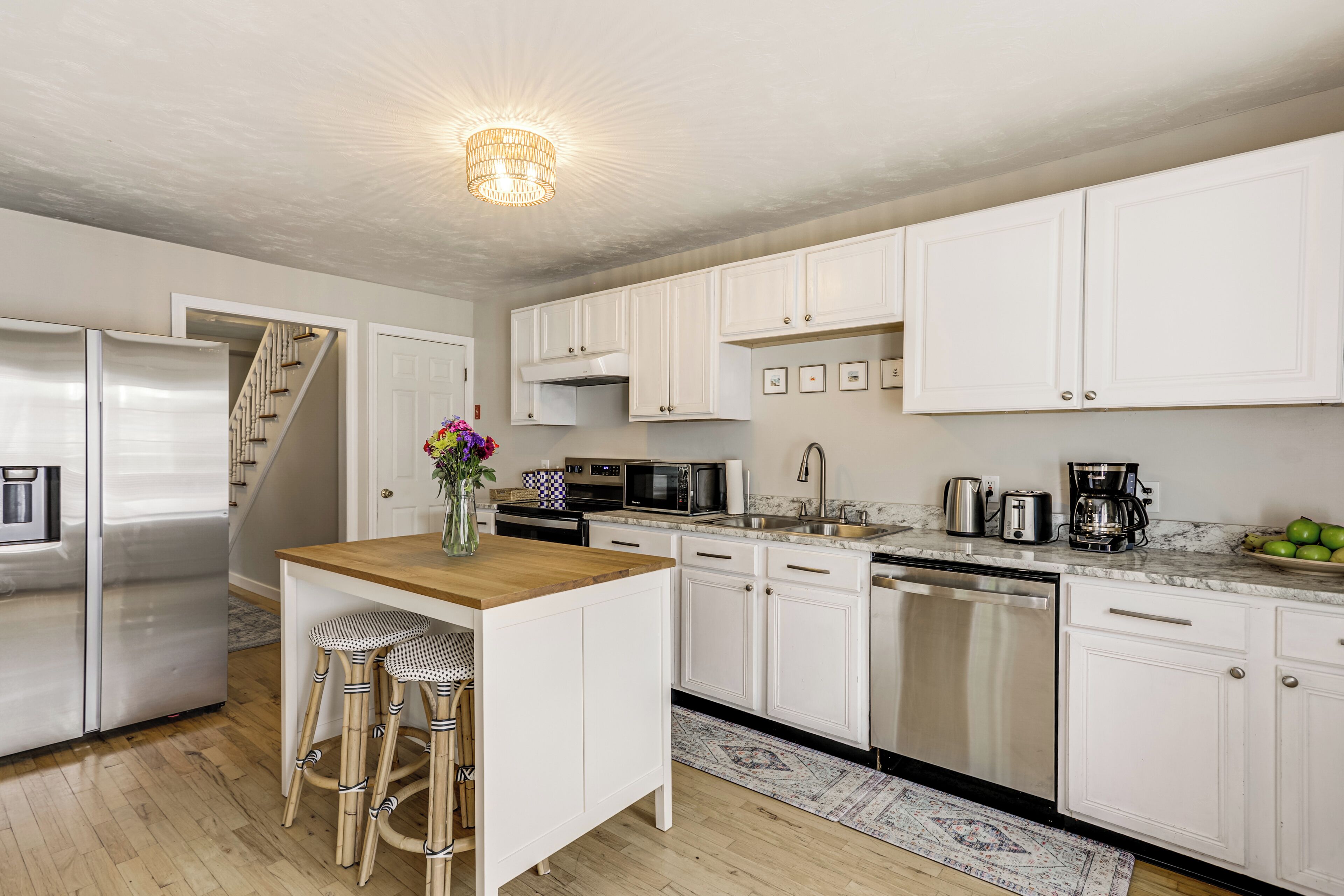A modern kitchen with white cabinets, stainless steel appliances, a wooden island with stools, and a patterned rug.
