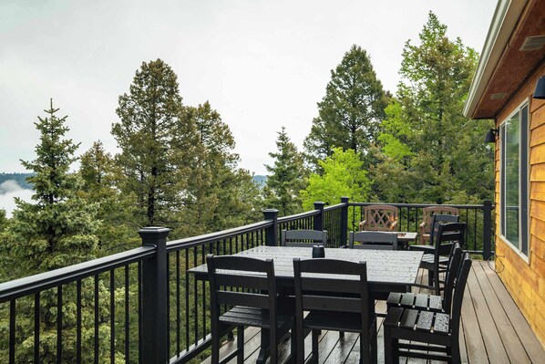 A wooden deck with a table and chairs, surrounded by a black railing, overlooking a forested area.