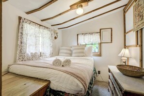 Une chambre avec un plafond en bois, un lit avec des serviettes roulées, une fenêtre avec des rideaux et une table de chevet avec une lampe et un panier.