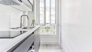Eine schmale Küche mit weißen Fliesen, einer Edelstahl-Spüle und einem Fenster mit dekorativem Gitter.