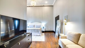 A modern bedroom with a large flat-screen TV, a bed with white bedding, a beige sofa, and a chandelier. - Historic Downtown Marshall: Efficient Studio Space (Marshall)