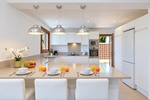A modern kitchen with a central island, white cabinetry, stainless steel appliances, pendant lighting, and a breakfast setup with fruit and orange juice.