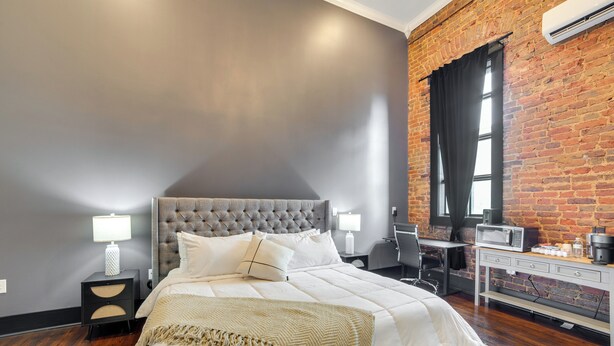 Un dormitorio moderno con un cabecero tapizado en gris, una cama grande con ropa de cama blanca, una mesita de noche con una lámpara, un escritorio con una computadora y una pared de ladrillo.