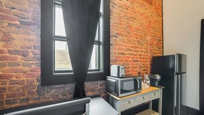 Una cocina compacta con pared de ladrillo, una ventana con cortinas negras, microondas, refrigerador y una mesita.