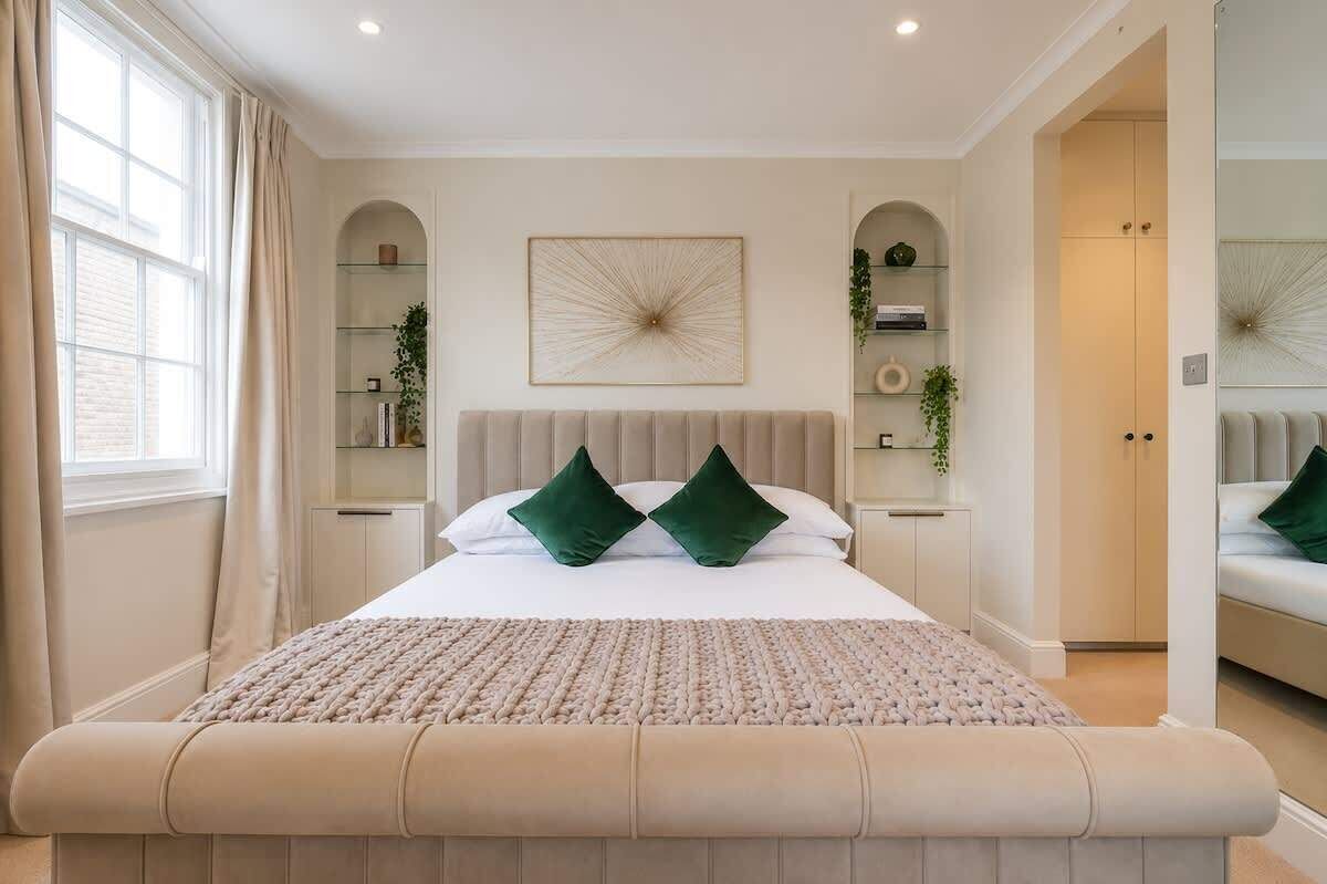 A bedroom with a large bed, a bench at the foot of the bed, a window with curtains, and built-in shelving with plants.