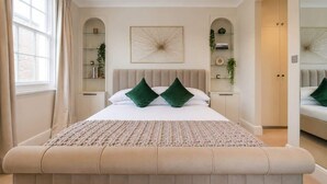 Une chambre avec un grand lit, un banc au pied du lit, une fenêtre avec des rideaux et des étagères intégrées ornées de plantes.
