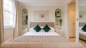 A bedroom with a large bed, a bench at the foot of the bed, a window with curtains, and built-in shelving with plants.