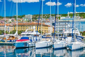 A marina with multiple sailboats docked, a cityscape with buildings in the background, and a clear blue sky. - Nurli Apt Near Glavanovo Beach (Rijeka)
