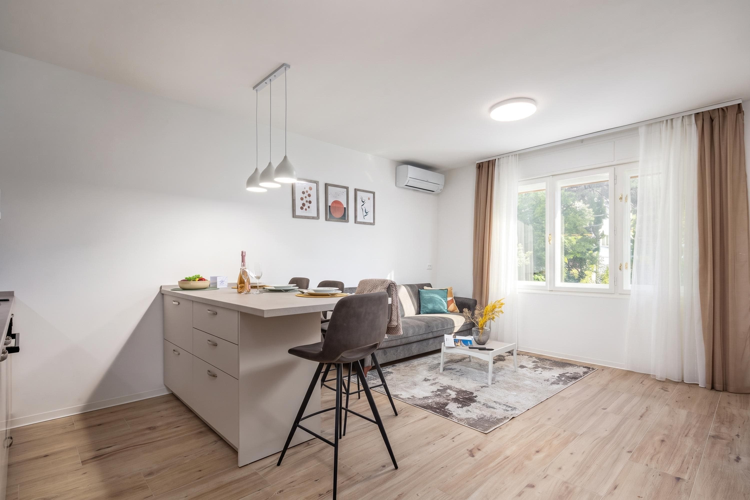 A modern kitchen with a dining area, a built-in island, pendant lighting, and a view through a large window.